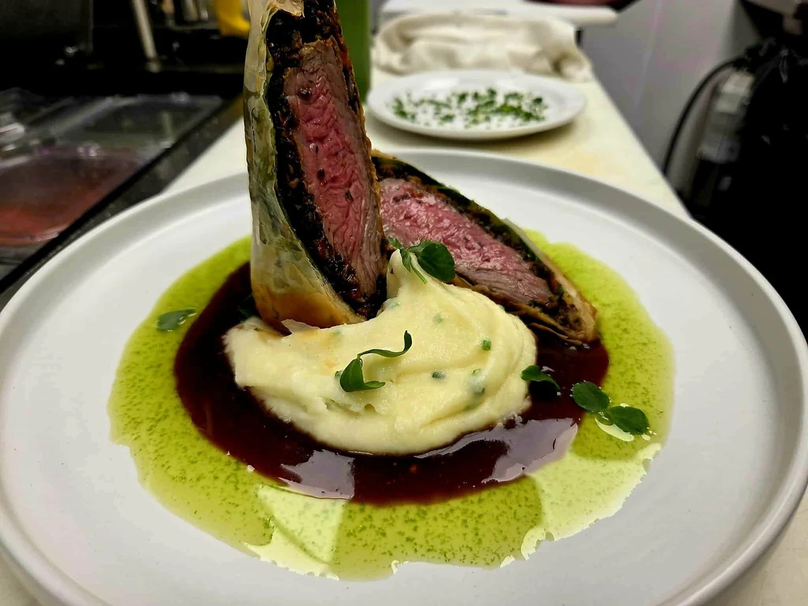 Sliced pastry-wrapped meat with mashed potato and green and dark sauces on a white plate